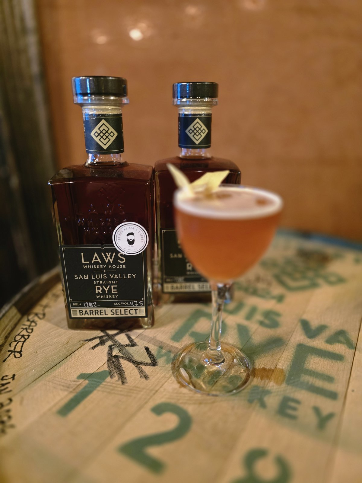 Two bottles of Laws Whiskey House San Luis Valley Straight Rye — Barrel Select for The Bookcase and Barber — beside a rye cocktail on the custom whiskey barrel